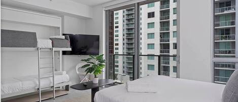 A modern bedroom with a bed, a TV, a plant, and a view of a high-rise building.