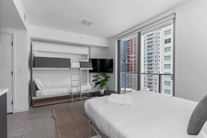 A modern bedroom with a bunk bed, a flat-screen TV, a potted plant, and a large window with a city view. - NEW Lux Condo - High-Floor Views · Gym · Pool · (Miami)