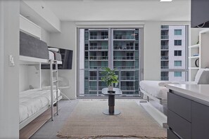Habitació moderna i ben il·luminada amb un llit superposat, una taula petita amb una planta i un gran finestral amb vista a un edifici.