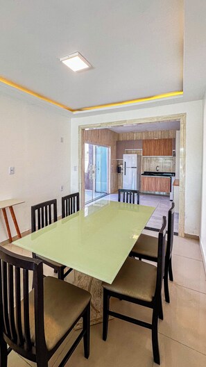 A dining area with a rectangular table and six chairs, a kitchen in the background, and a ceiling light fixture. - Casa Summer Ville em Barreirinhas/em condomínio/piscina privativa (Barreirinhas)