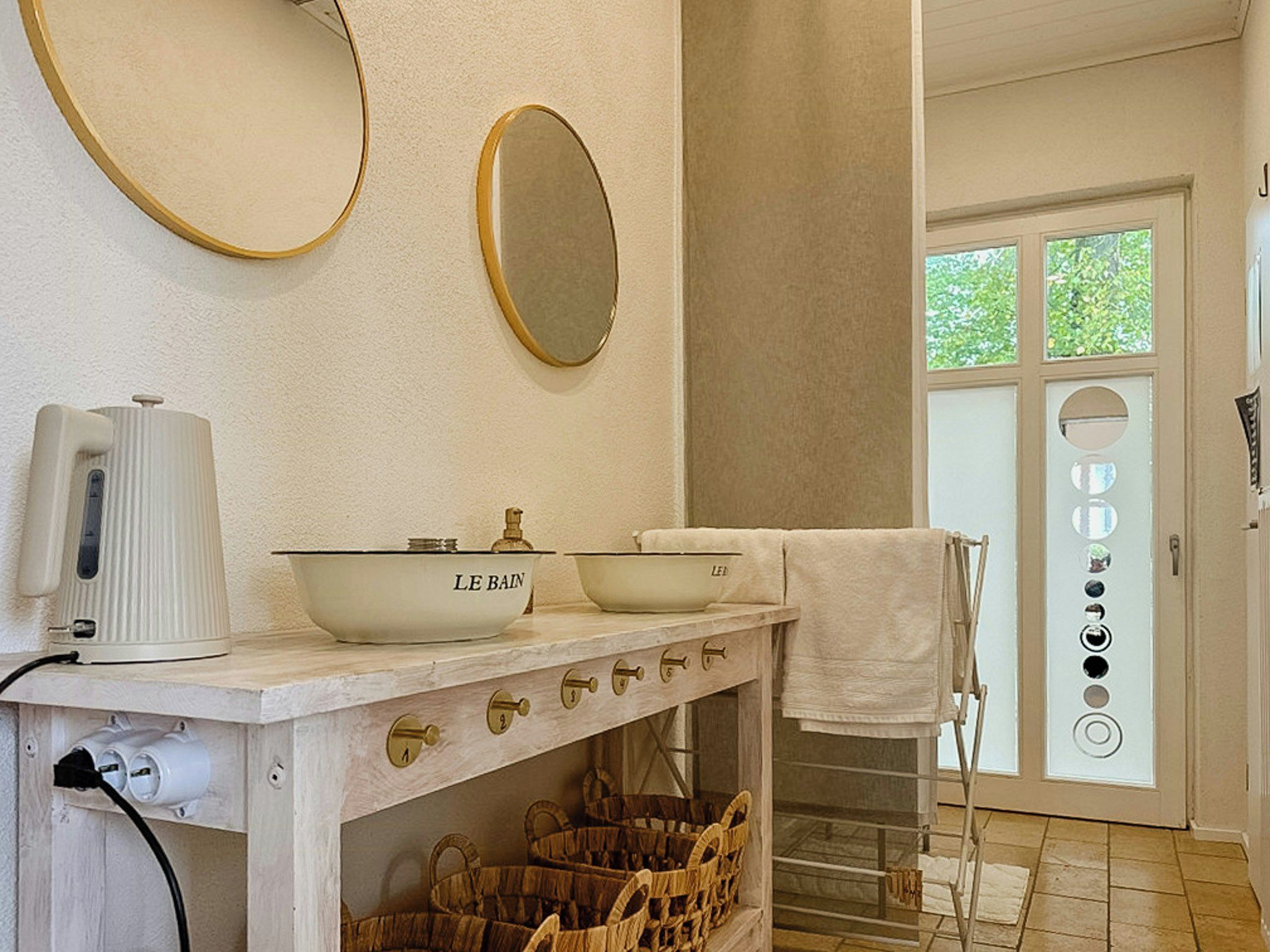A bathroom with a white sink, two bowls labeled 'LE BAIN,' a white towel, a mirror, and a window with a view of greenery.