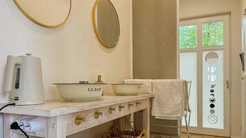 A bathroom with a white sink, two bowls labeled 'LE BAIN,' a white towel, a mirror, and a window with a view of greenery.