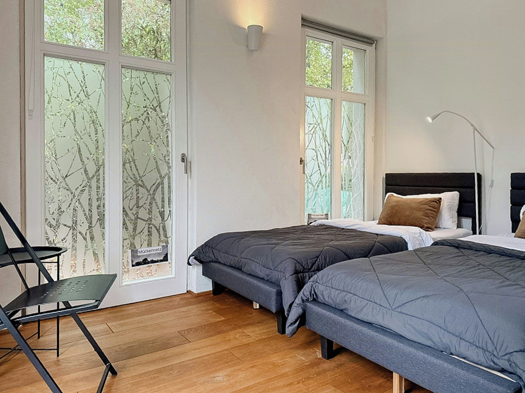 A bedroom with two beds, a chair, and large windows with a view of trees.