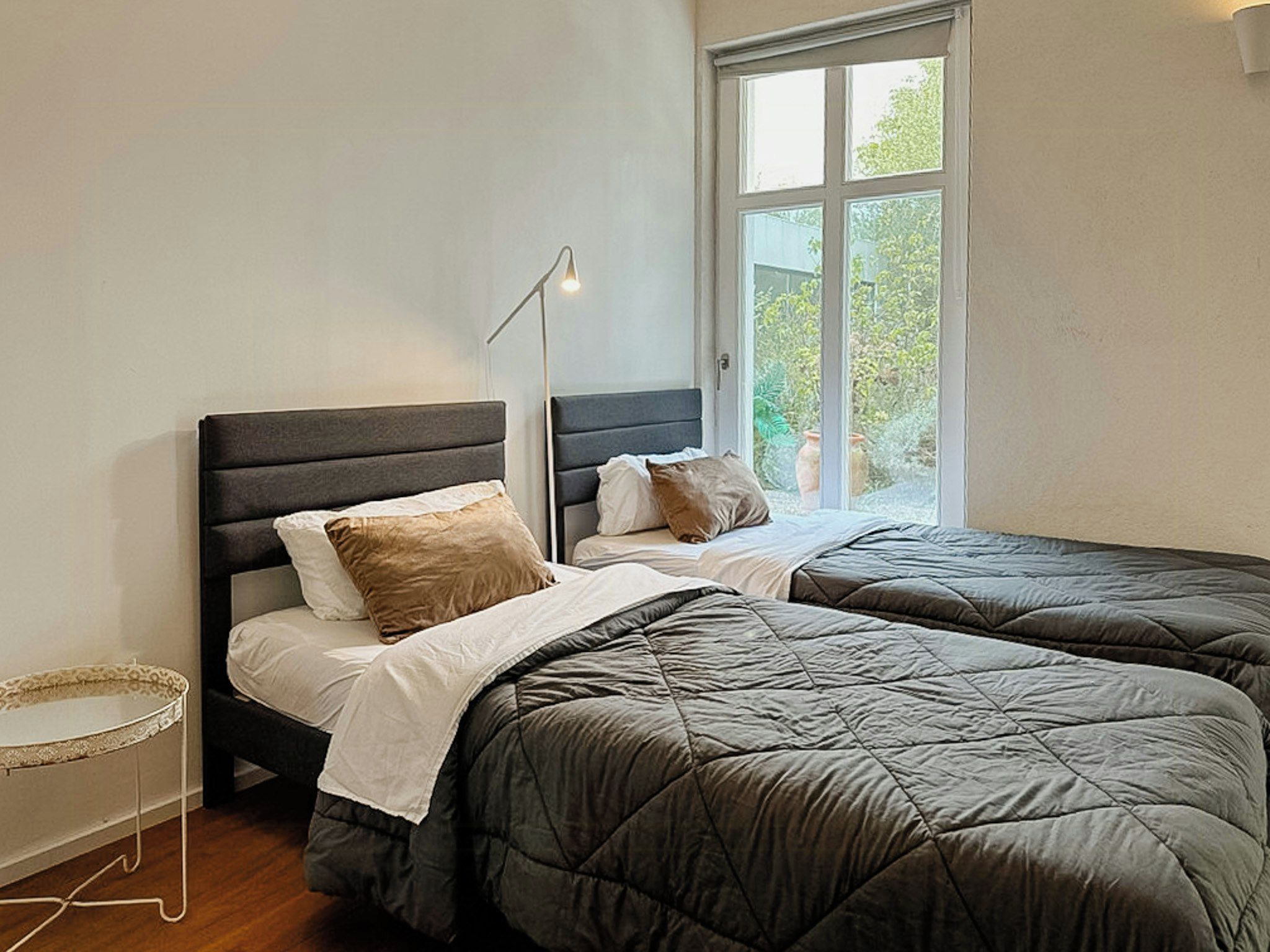 A bedroom with two beds, a bedside table, and a window with a view of greenery.