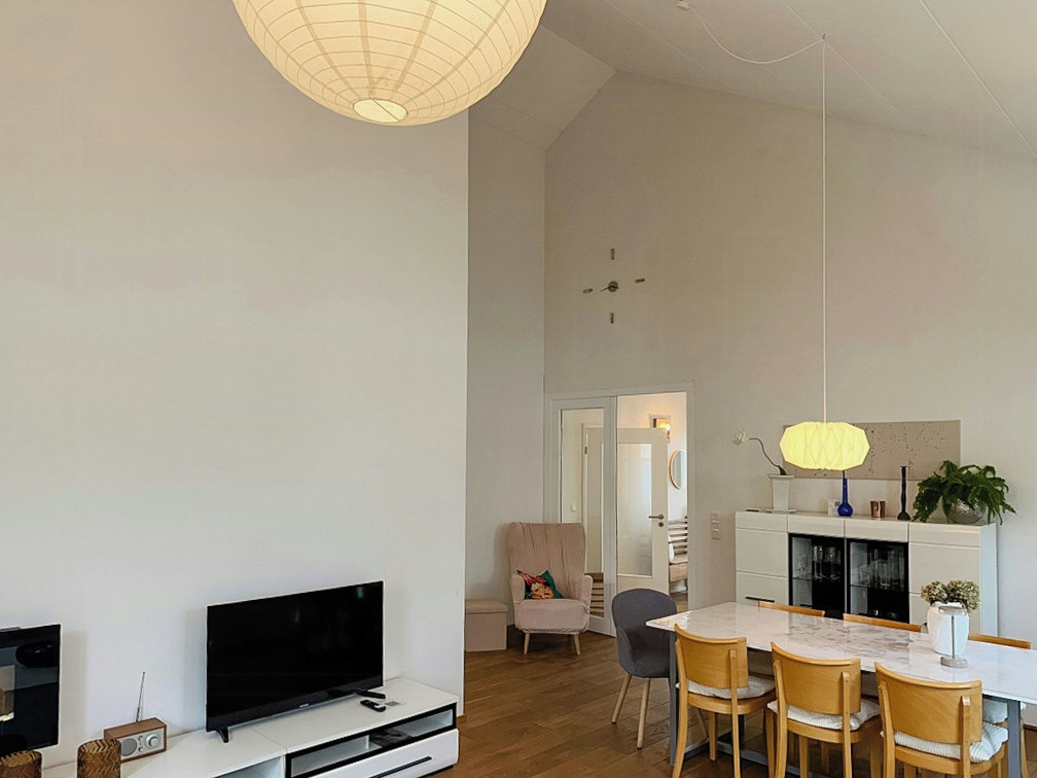 Una sala moderna con un televisor de pantalla plana, una mesa de comedor con sillas, y una cocina con armarios y una lámpara colgante.