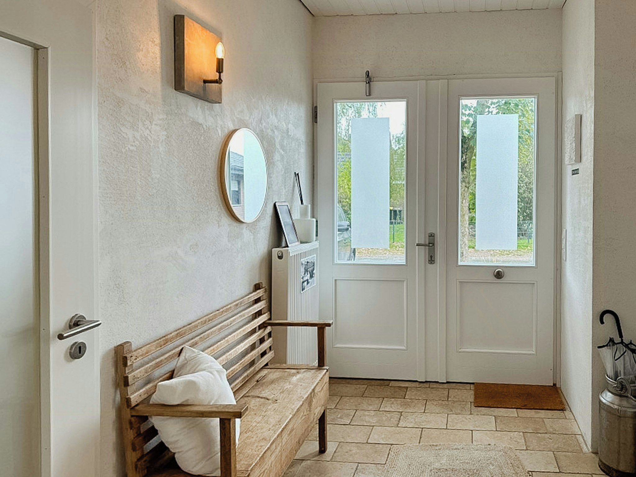 A room with a bench, a mirror, a wall-mounted light, and a door with a view of the outdoors.