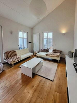 A modern living room with a white sofa, a white coffee table, and a wooden floor. There are two windows with curtains, a lamp, and a small statue on a table.