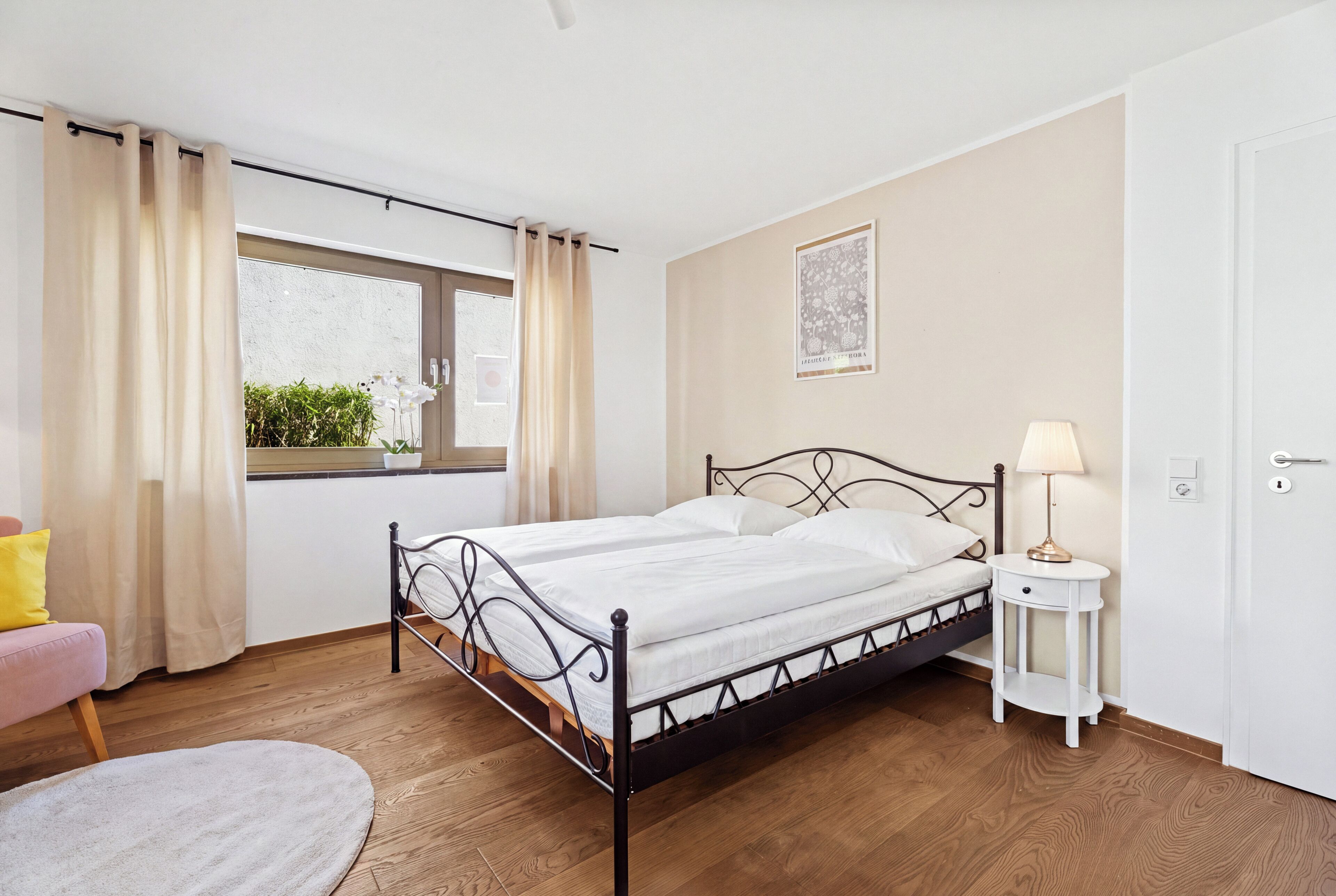 Ein Einzelbett mit Metallgestell, ein Nachttisch mit Lampe, ein Fenster mit Vorhängen und ein kleiner Topfpflanze am Fensterbrett.