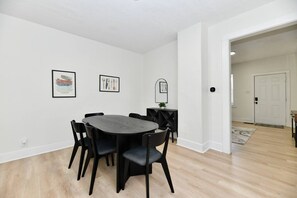 Un coin repas avec une table ronde et des chaises, des murs blancs et une porte donnant sur une autre pièce.