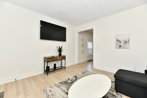 Un salon moderne avec un téléviseur à écran plat, une console en bois agrémentée d’une plante, un fauteuil blanc et un canapé noir.