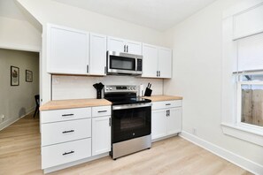 A compact kitchen with white cabinetry, a microwave, a stove, and a window with blinds. - Fenced yard • pets • parking • IMS + downtown (Indianapolis)