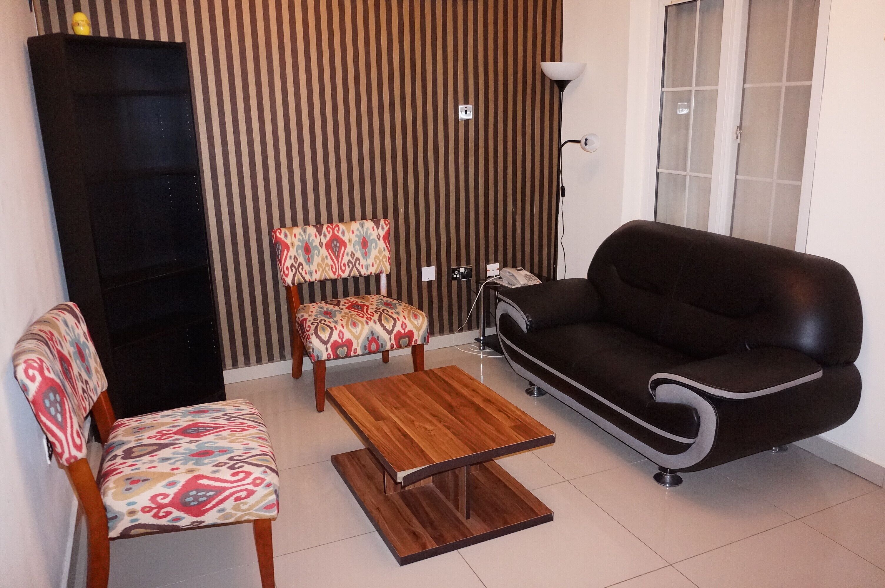 Uma sala de estar com um sofá, duas cadeiras estampadas, uma mesa de centro de madeira, um armário preto, um abajur e um telefone.