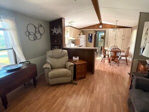 Ein gemütliches Wohnzimmer mit Ledersessel, Esstisch aus Holz und Küchenzeile mit weißen Schränken und Holzboden.