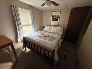 Ein Schlafzimmer mit einem Bett, einem Deckenventilator, einem Fenster mit Vorhängen, einem Nachttisch und einem gerahmten Bild an der Wand.