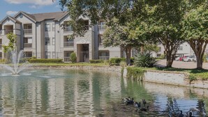 A multi-story residential building with a central courtyard featuring a fountain and a pond with ducks. - Landing | Stylish 1BD, Gym, Clubhouse (Houston)