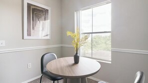 A room with a round table, a chair, a vase with flowers, a framed picture on the wall, and a window with a view. - Landing | Modern 1BD, Gym, Clubhouse (Houston)