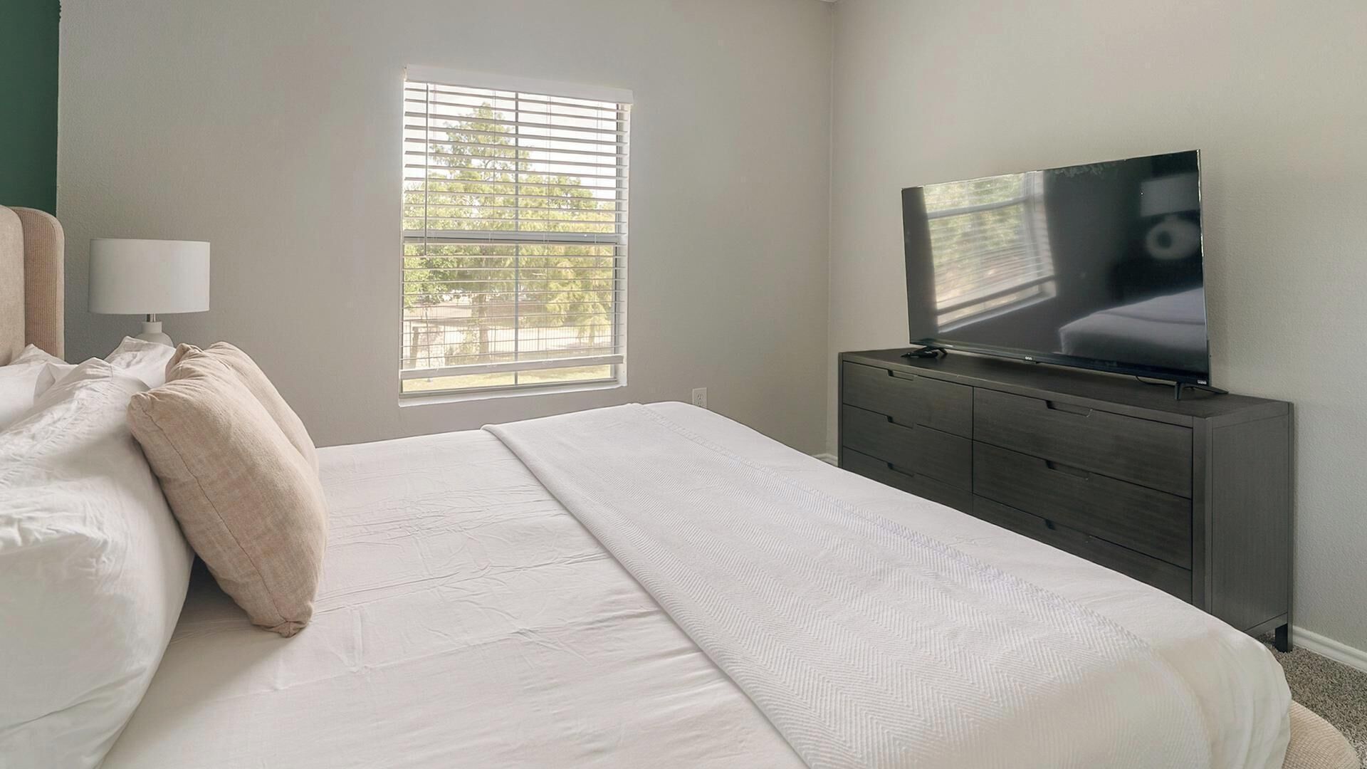 Une chambre à coucher comprenant un lit, une commode, un téléviseur et une fenêtre avec des stores.