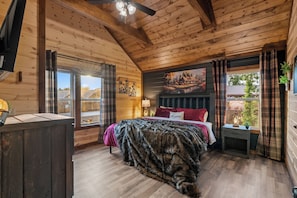 Ein Schlafzimmer in einer Holzhütte mit einem Bett, einem Fenster mit Vorhängen, einem Bild an der Wand und einem Deckenventilator.
