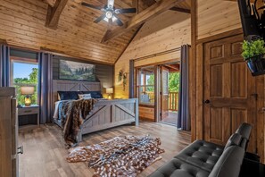 A wooden cabin interior with a bed, a desk, a chair, a ceiling fan, and a view of the outdoors. - Luxury BRAND NEW 8bdrm Cabin-Pool-Arcades-Gazebo (Sevierville)