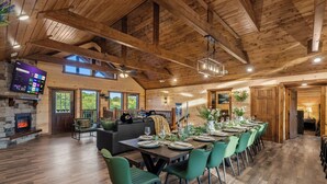A spacious room with a high ceiling, wooden beams, a fireplace, a large flat-screen TV, and a dining area set for a meal. - Luxury BRAND NEW 8bdrm Cabin-Pool-Arcades-Gazebo (Sevierville)