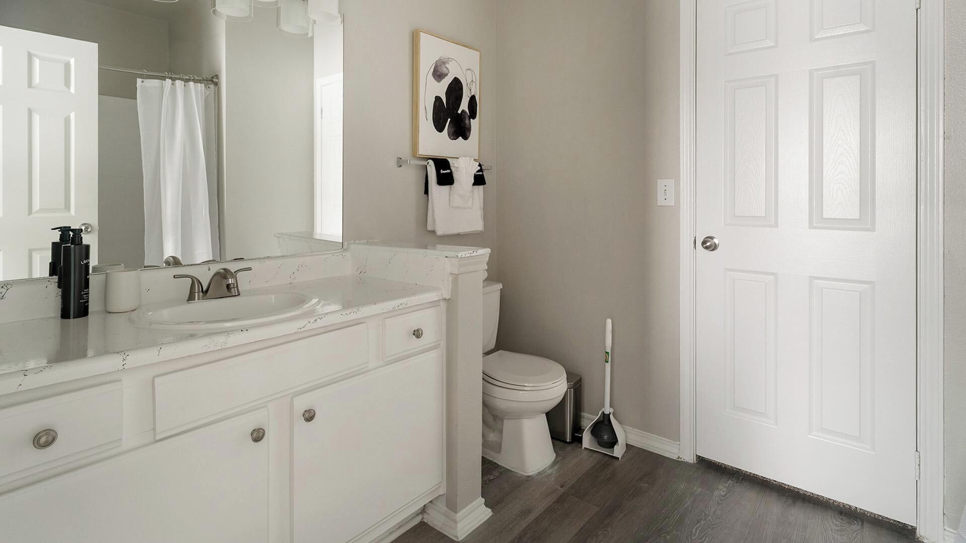 A bathroom with a white vanity, sink, and toilet. There is a painting on the wall and a broom leaning against the wall.