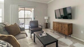 A living room with a sofa, armchair, coffee table, TV, and a sliding glass door leading to an outdoor area. - Landing | Stunning 1BD, Clubhouse, Gym (Houston)