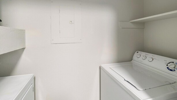 A laundry room with a white washing machine and dryer, a wall-mounted electrical outlet, and a shelf above the dryer. - Landing | Amazing 2BD, Gym, Clubhouse (Houston)