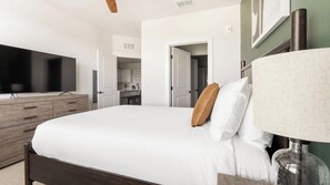 Ein Schlafzimmer mit einem großen Bett, einem Fernseher, einer Holzkommode und einer durch eine offene Tür einsehbaren Küchenzeile.