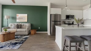 A modern kitchen with a stainless steel refrigerator, a microwave, and a stove. There is a dining area with a bar stool and a coffee table. - Landing | Amazing 1BD, Gym, Game Room (Punta Gorda)