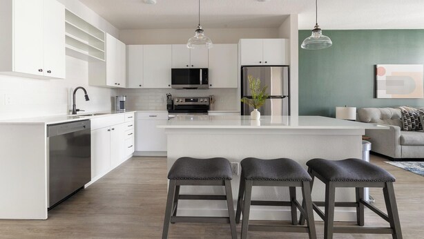 Una cocina moderna con armarios blancos, una isla central con taburetes y una zona de comedor con sofá.