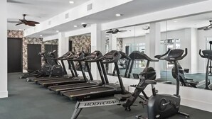 Un gimnasio con cintas de correr y una máquina elíptica, paredes blancas y un ventilador de techo.