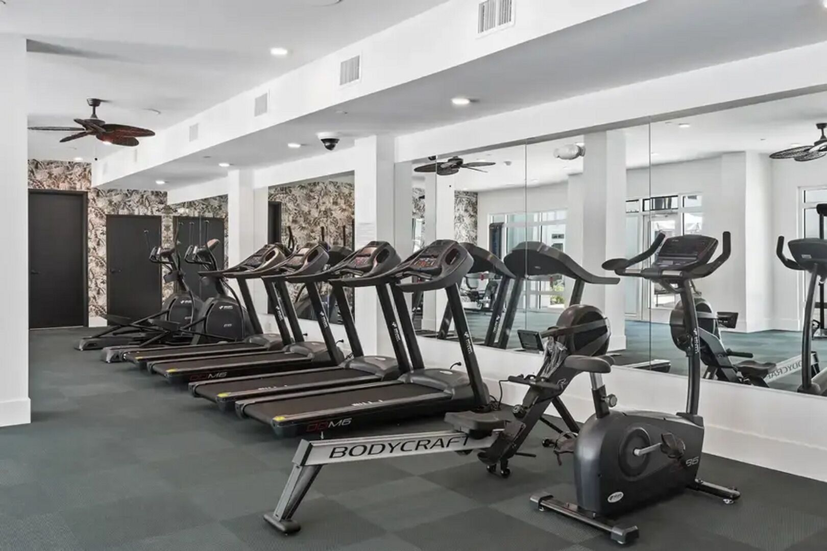 A gym with multiple treadmills and an elliptical machine, white walls, and a ceiling fan.