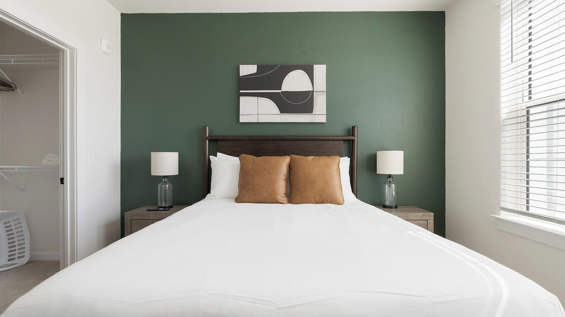A hotel room with a bed, two bedside lamps, a wall art piece, and a window with blinds.