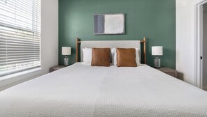 Un lit bien fait, recouvert d’une couvre-lit blanche à texture, et agrémenté de deux oreillers marron, est placé de part et d’autre de deux tables de chevet sur lesquelles reposent des lampes. La pièce est dotée de murs verts et d’une fenêtre équipée de stores.