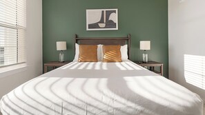 A neatly made bed with a white comforter and two brown pillows, flanked by nightstands with lamps, in a room with green walls and a framed artwork above the bed. - Landing | Spectacular 2BD, Gym, Game Room (Punta Gorda)