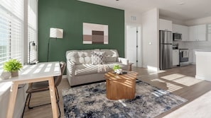 A modern living room with a green accent wall, a sofa, a coffee table, and a kitchen area with a refrigerator and microwave. - Landing | Spectacular 2BD, Gym, Game Room (Punta Gorda)