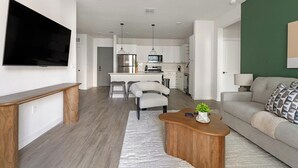 A modern living room with a flat-screen TV, a wooden coffee table, a grey sofa, and a kitchen in the background. - Landing | Stunning 2BD, Game Room, Gym (Punta Gorda)