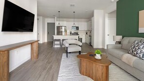 Ein modernes Wohnzimmer mit Flachbildfernseher, Couchtisch aus Holz, grauem Sofa und einer Küche im Hintergrund.