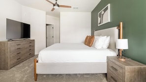 Quarto com cama, televisão, penteadeira, abajur e ventoinha no teto.