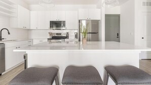 Een moderne keuken met witte kasten, roestvrijstalen apparatuur en een centraal eiland. Voor het eiland staan drie gestoffeerde grijze krukken.