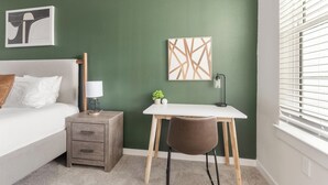 Een slaapkamer met een bed, nachtkastje, lamp, bureau, stoel en een raam met jaloezieën.