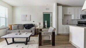 A modern living room with a sofa, coffee table, and a chair. There is a kitchen area with cabinets and appliances. - Landing | Modern 2BD, Pool, Gym (Port Charlotte)