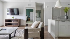 A modern living room with a flat-screen TV, a sofa, a coffee table, and a kitchen area with cabinets and a countertop. - Landing | Incredible 2BD, Gym, Pool (Port Charlotte)