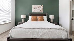 Ein ordentlich gemachtes Bett mit weißer Bettdecke und zwei braunen Kissen, flankiert von zwei Nachttischen mit Lampen, in einem Zimmer mit grüner Akzentwand und einem gerahmten abstrakten Gemälde über dem Bett.