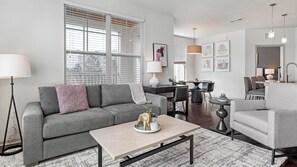 A modern living room with a grey sofa, a coffee table, and a dining area with a table and chairs. - Landing | Modern 1BD, Pool, Gym (Port Charlotte)