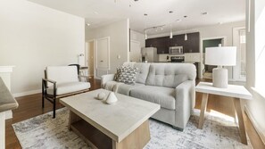 A modern living room with a sofa, a coffee table, and a dining area with a kitchen in the background. - Landing | Stylish 2BD, Gym, Pool (Austin)