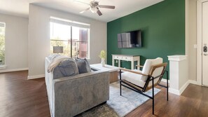 A modern living room with a grey sofa, a white armchair, a flat-screen TV mounted on a green wall, and a ceiling fan. - Landing | Spectacular 2BD, Pool, Gym (Austin)