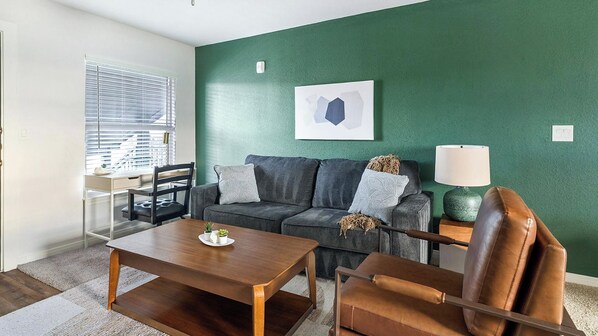 A living room with a dark green wall, a grey sofa with cushions, a wooden coffee table, a brown leather chair, a lamp, and a window with blinds. - Landing | Stylish 1BD, Pool (Houston)