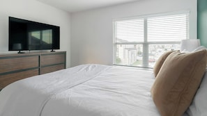 A hotel room with a bed, a TV on a wooden dresser, and a window with blinds. - Landing | Stylish 1BD, Pool (Houston)