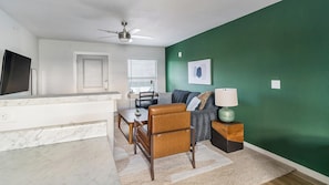 A modern living room with a green accent wall, a ceiling fan, a dining area with a table and chairs, and a kitchen counter. - Landing | Stylish 1BD, Pool (Houston)